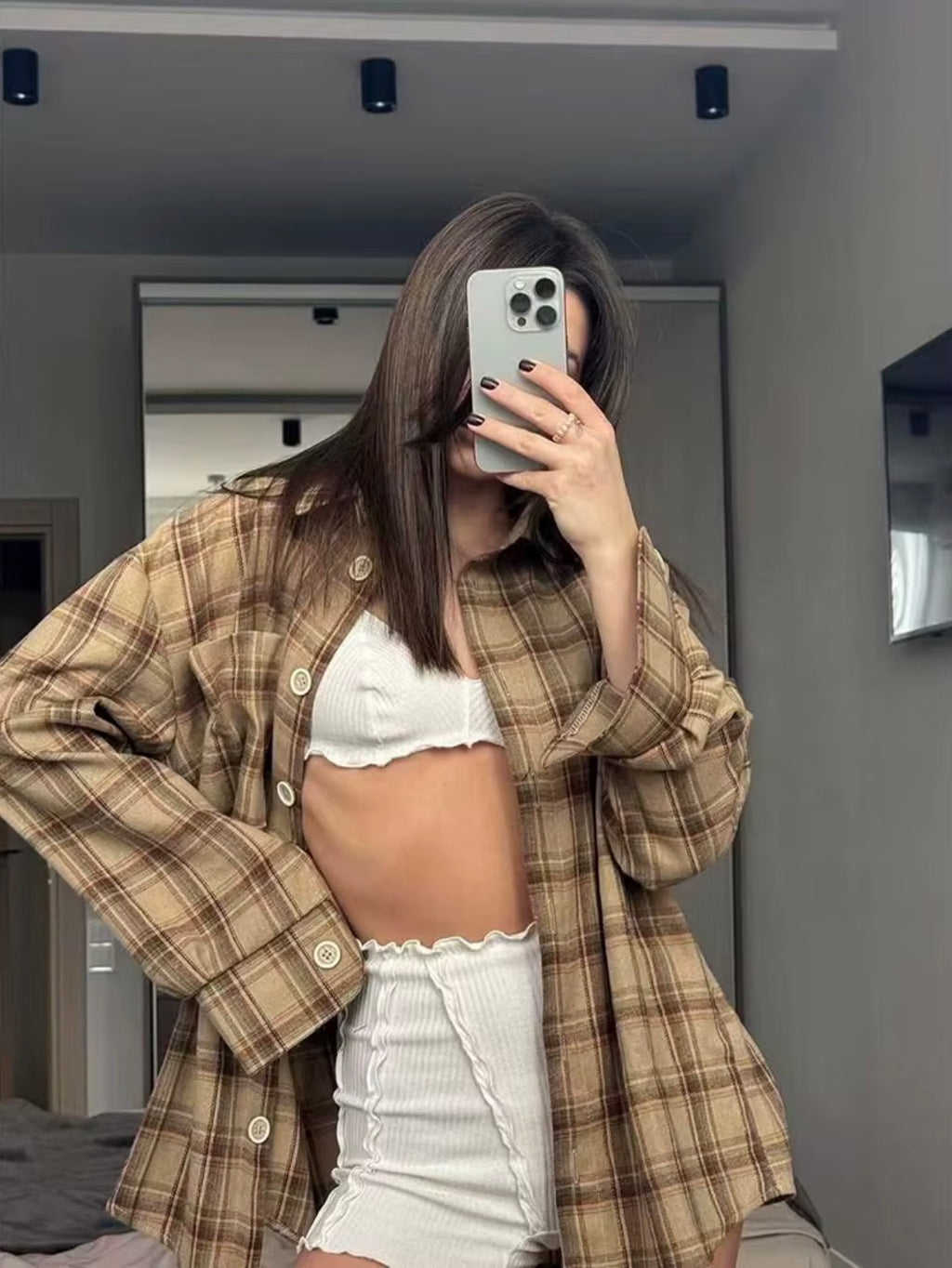 Person wearing a plaid shirt over a white top and shorts, taking a mirror selfie.