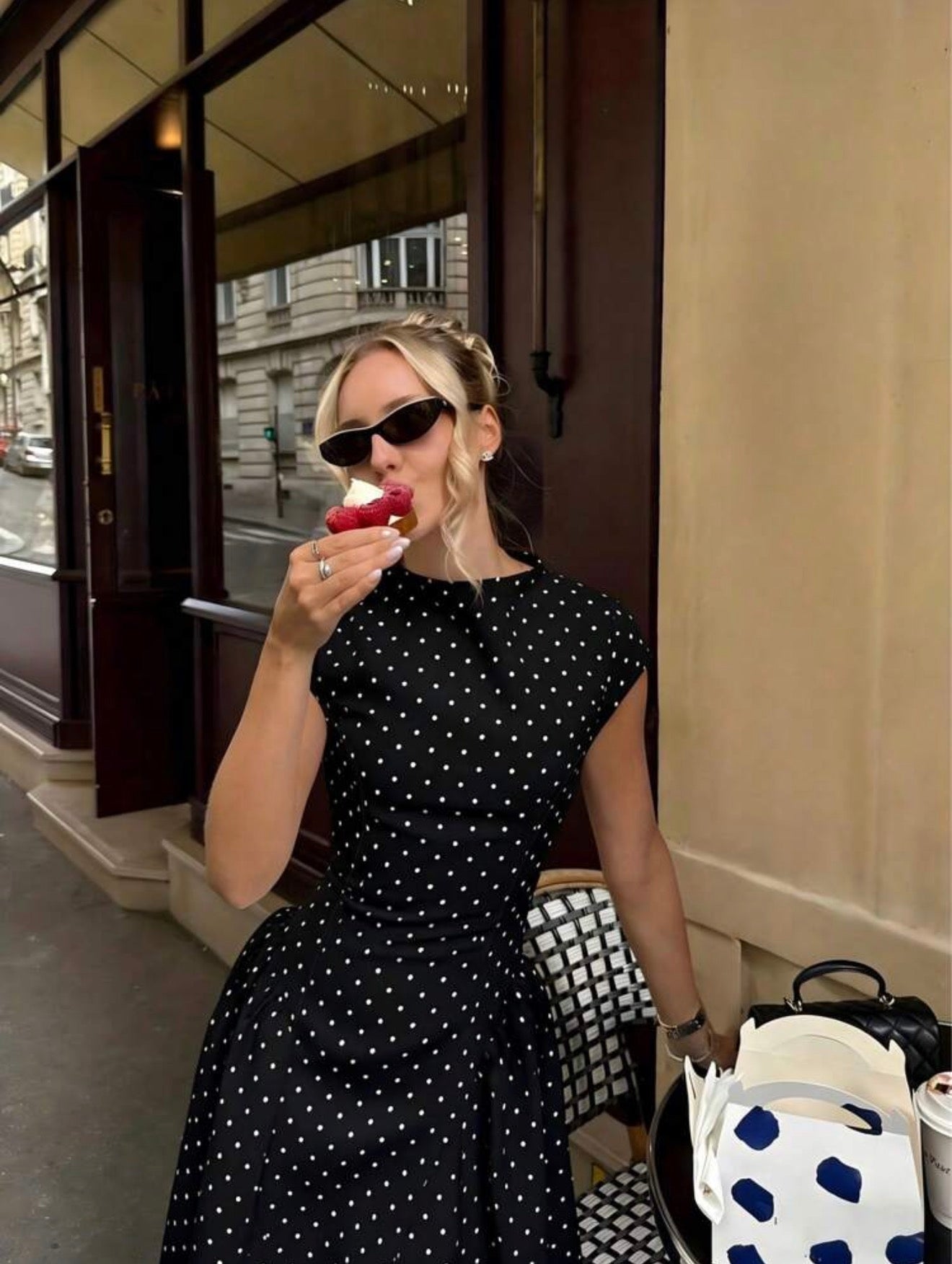 Woman in a black polka dot dress eating an apple outside a building.