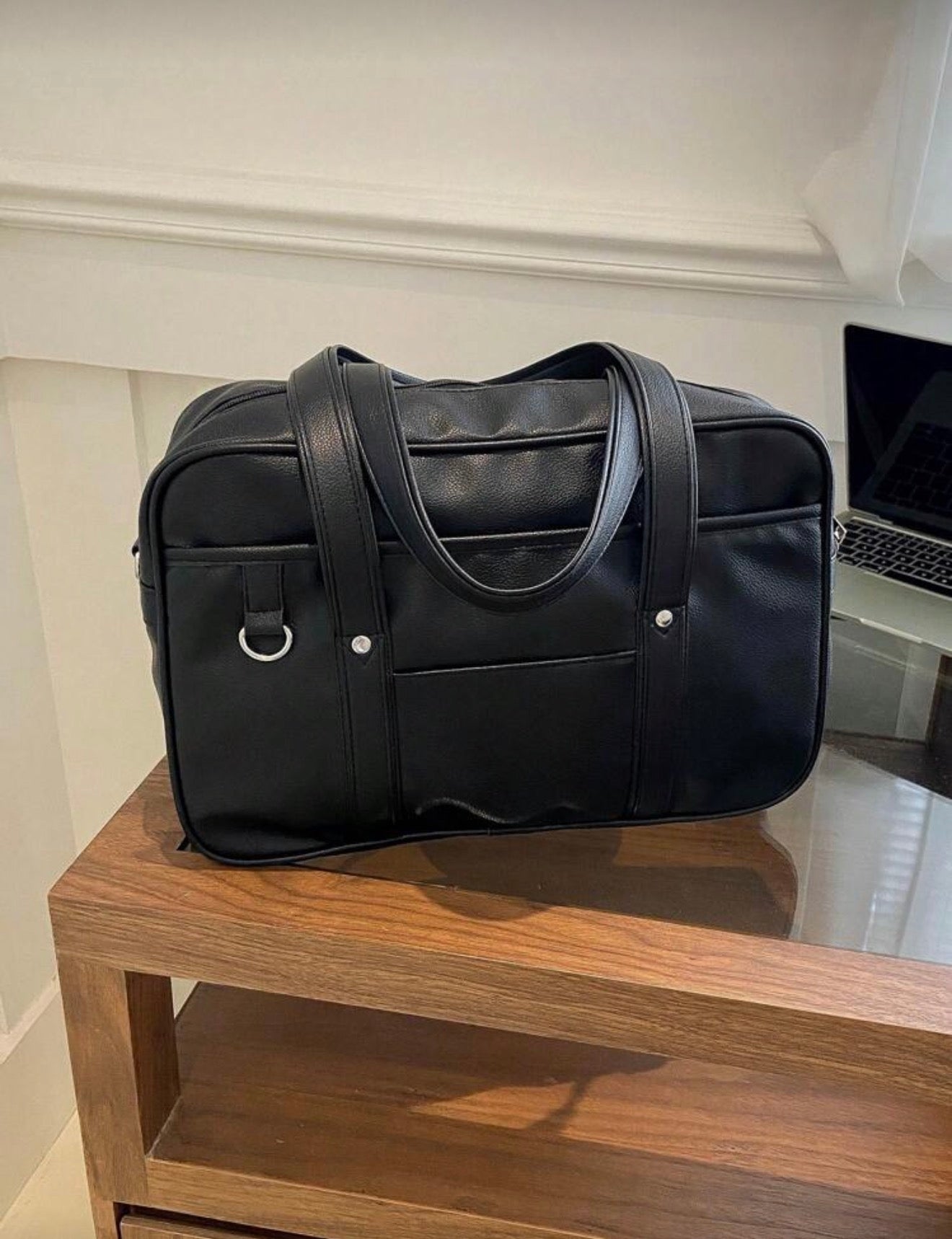 Black leather duffel bag on a wooden surface with a laptop in the background.