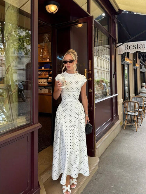 Woman in a white polka dot dress standing outside a restaurant holding a drink.