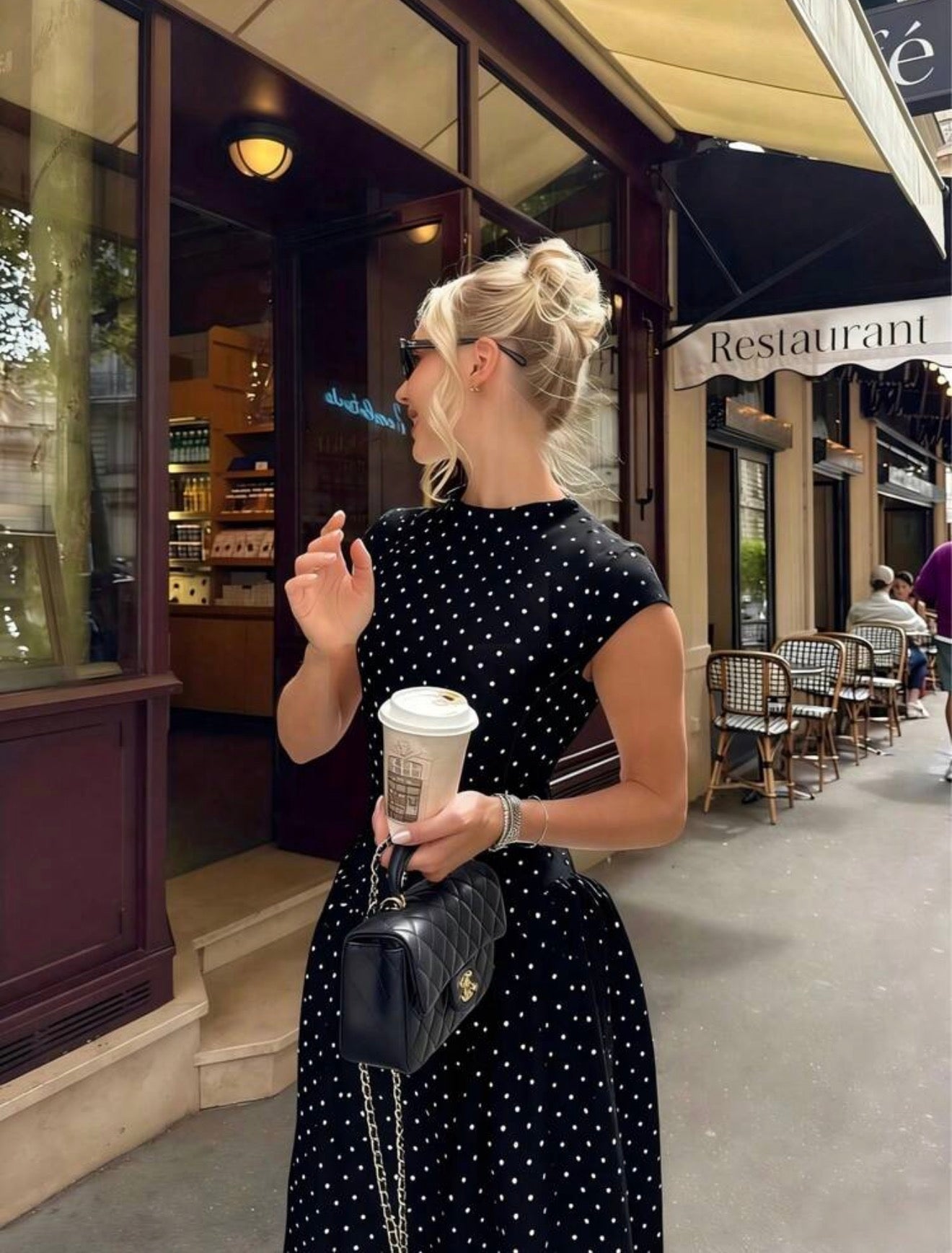 Woman in a polka dot dress holding a coffee cup and a handbag outside a restaurant.