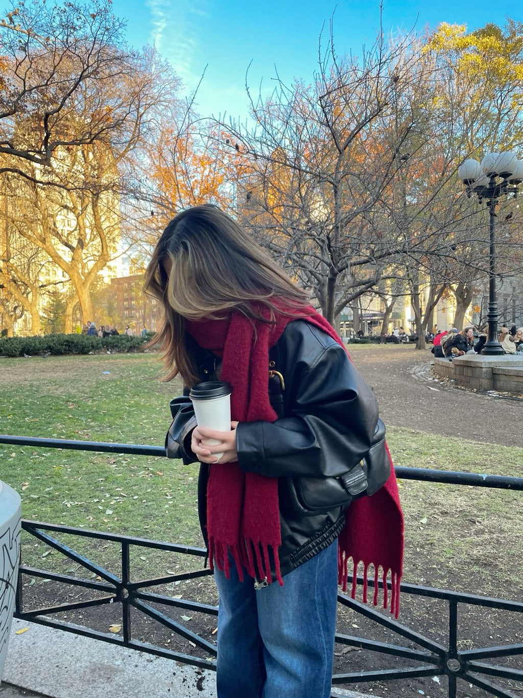 Person wearing a red scarf and black leather jacket in a park with autumn trees.