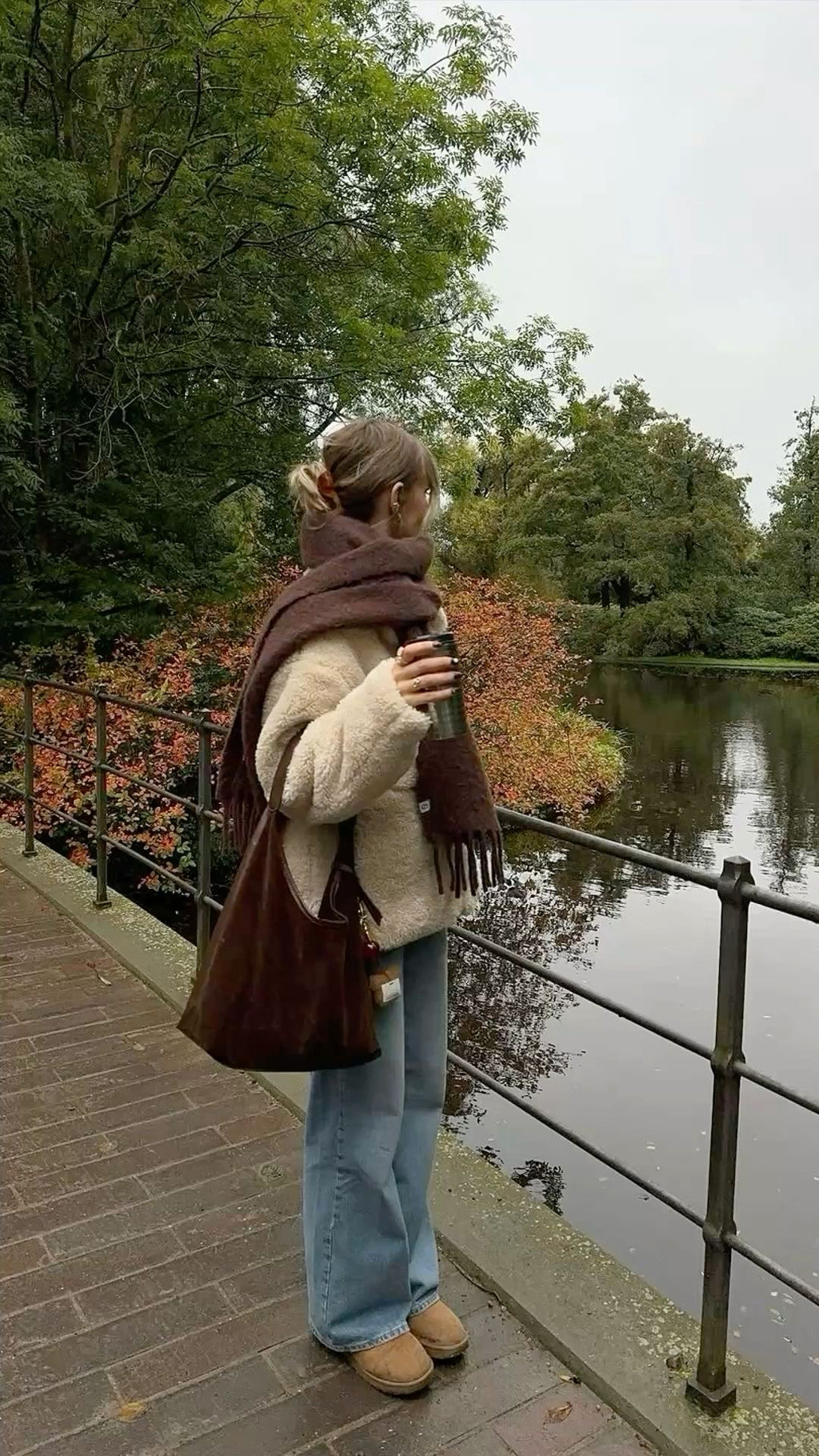 Person standing by a lake holding a coffee cup, surrounded by trees with autumn foliage.
