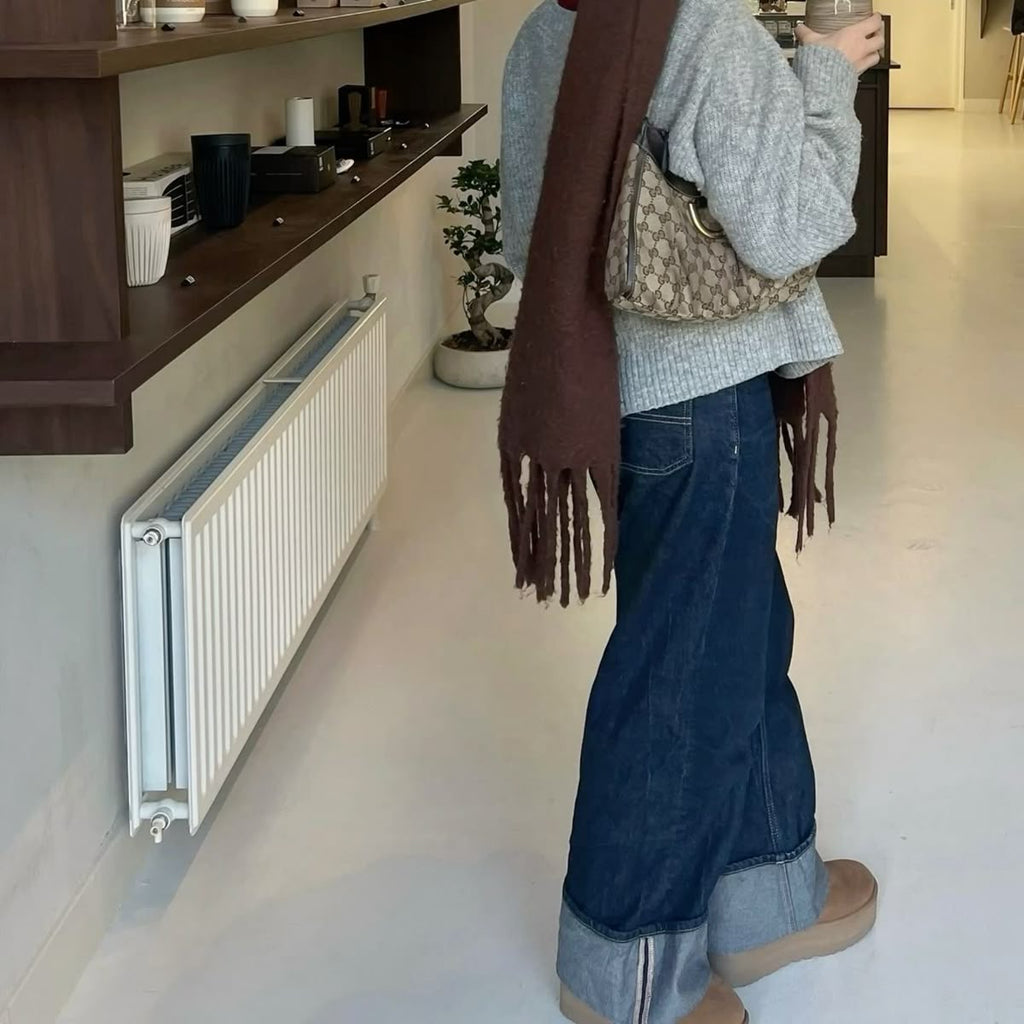 Person wearing a brown scarf and blue jeans standing in a room with a radiator and shelves.