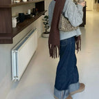 Person wearing a brown scarf and blue jeans standing in a room with a radiator and shelves.