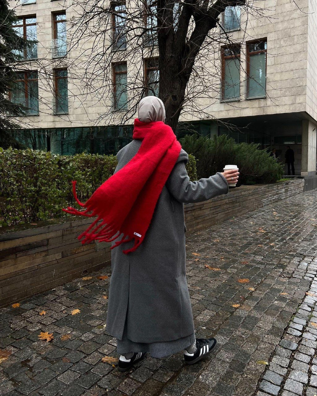 Person wearing a red scarf and gray coat walking on a sidewalk with a building in the background.