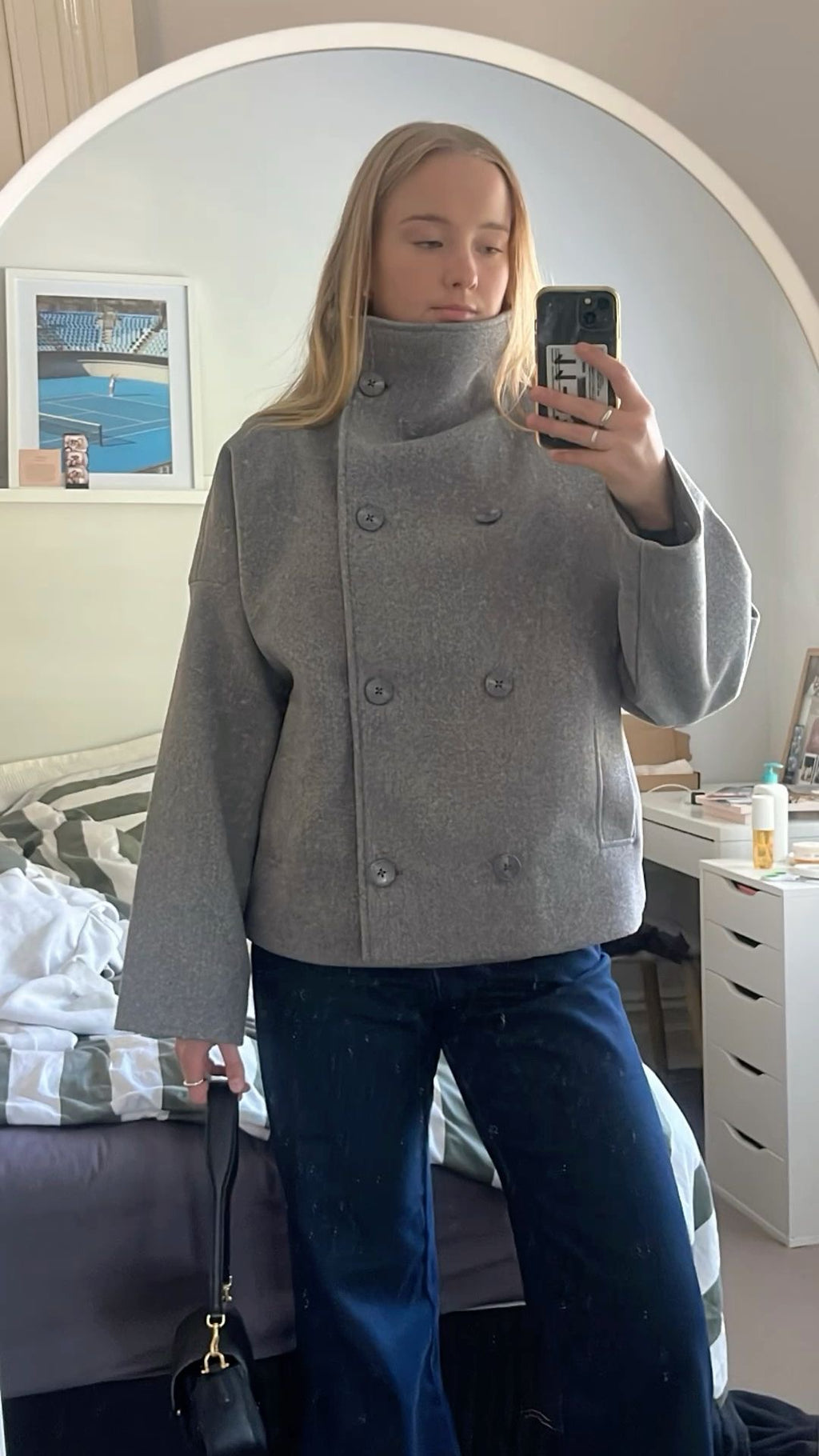 Person wearing a gray coat taking a mirror selfie in a bedroom.