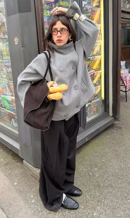 Person wearing a gray coat and black pants standing in front of a store with a bag and bread.