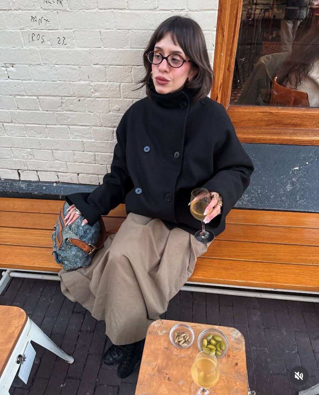 Woman sitting on a bench with a drink, holding a patterned clutch, in an urban setting.