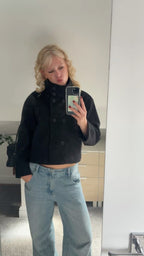 Person wearing a black coat and light blue jeans taking a mirror selfie.