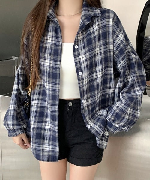 Person wearing a blue plaid shirt over a white top and black shorts indoors.