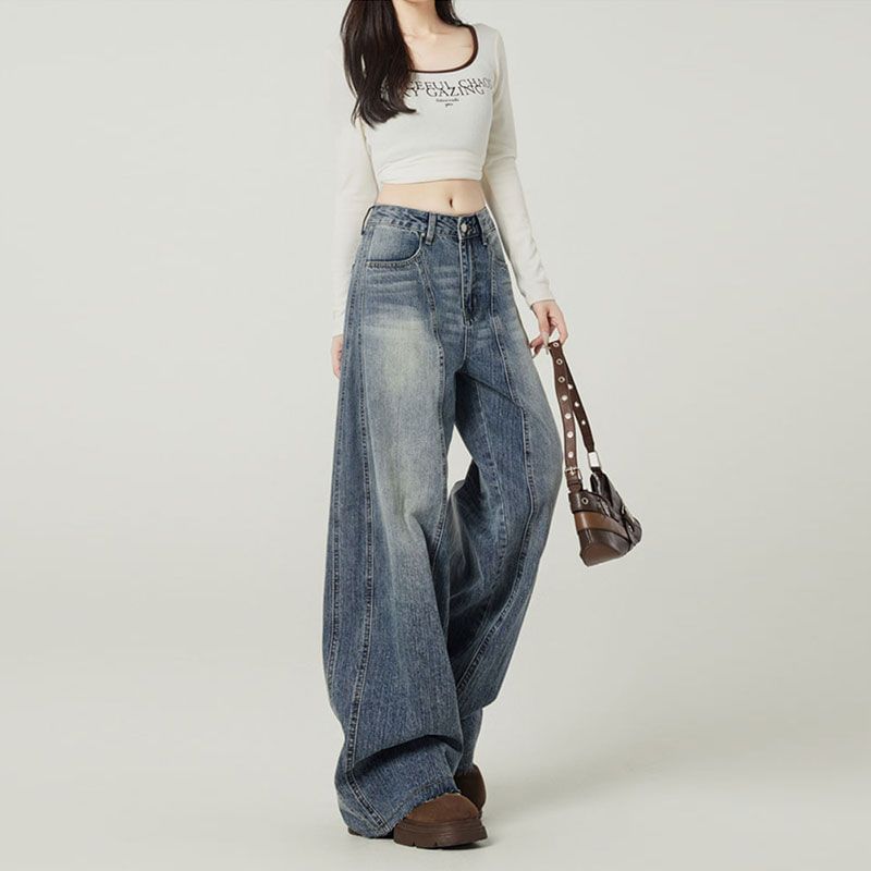Person wearing a white crop top and blue jeans holding a brown bag on a plain background