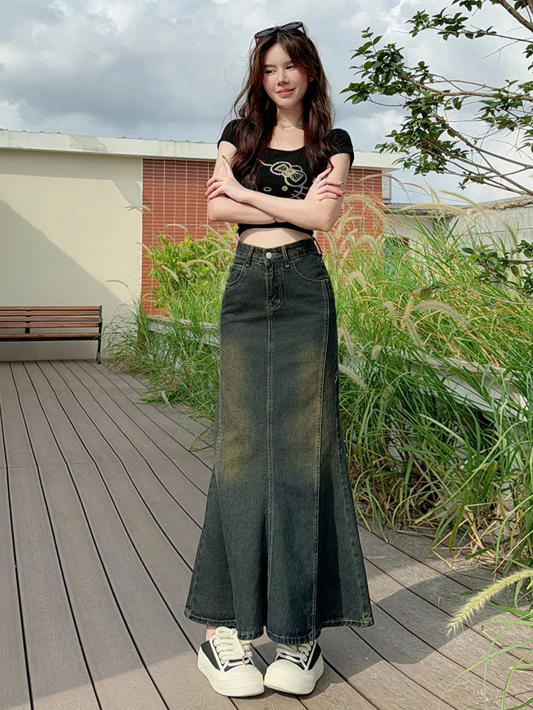 Woman wearing a black top and long denim skirt standing on a wooden deck with greenery in the background