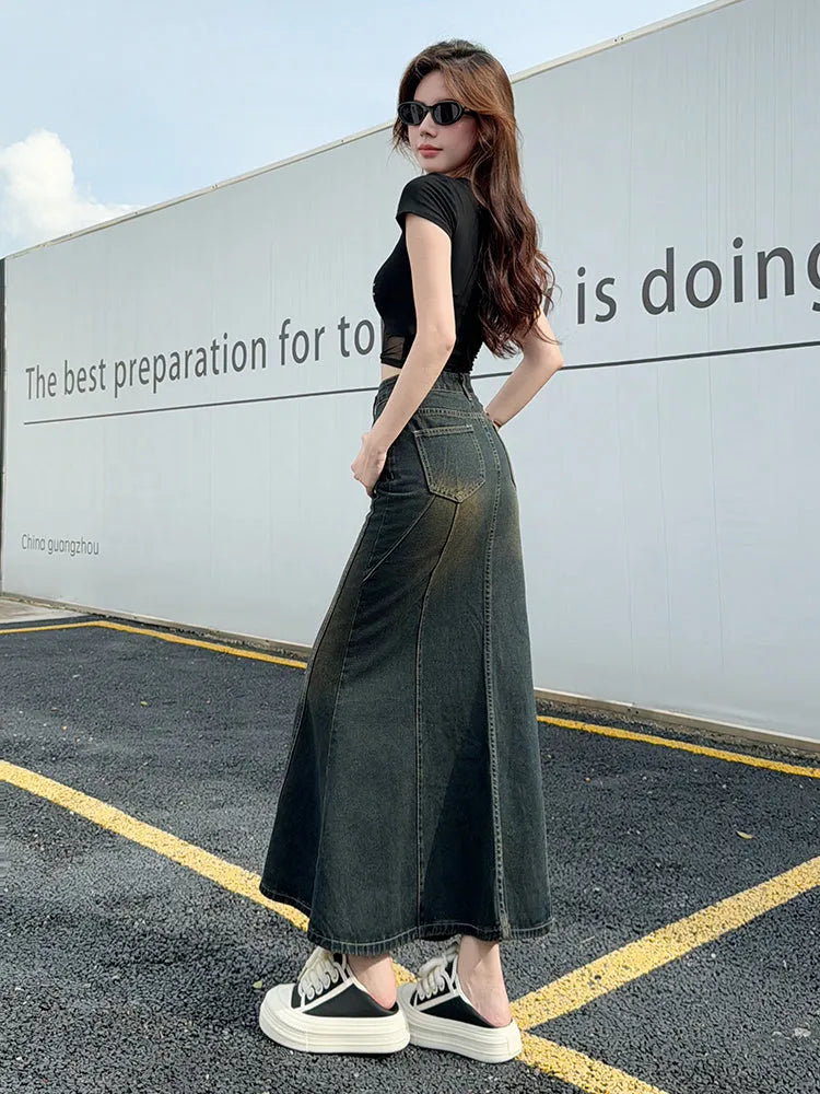 Woman wearing a black top and long denim skirt standing in front of a wall with text.