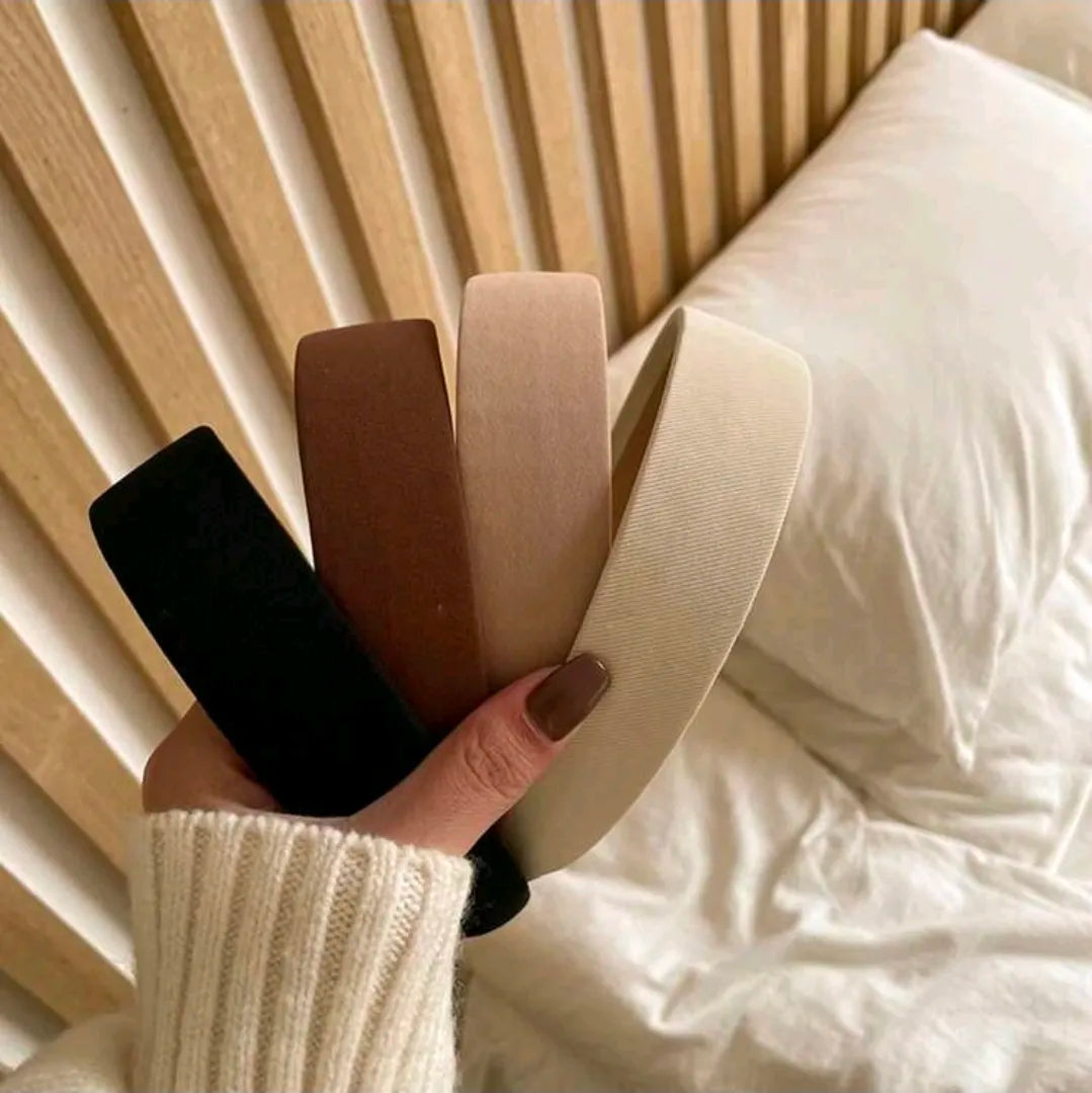 Hand holding four hairbands in black, brown, beige, and white against a blurred indoor background.