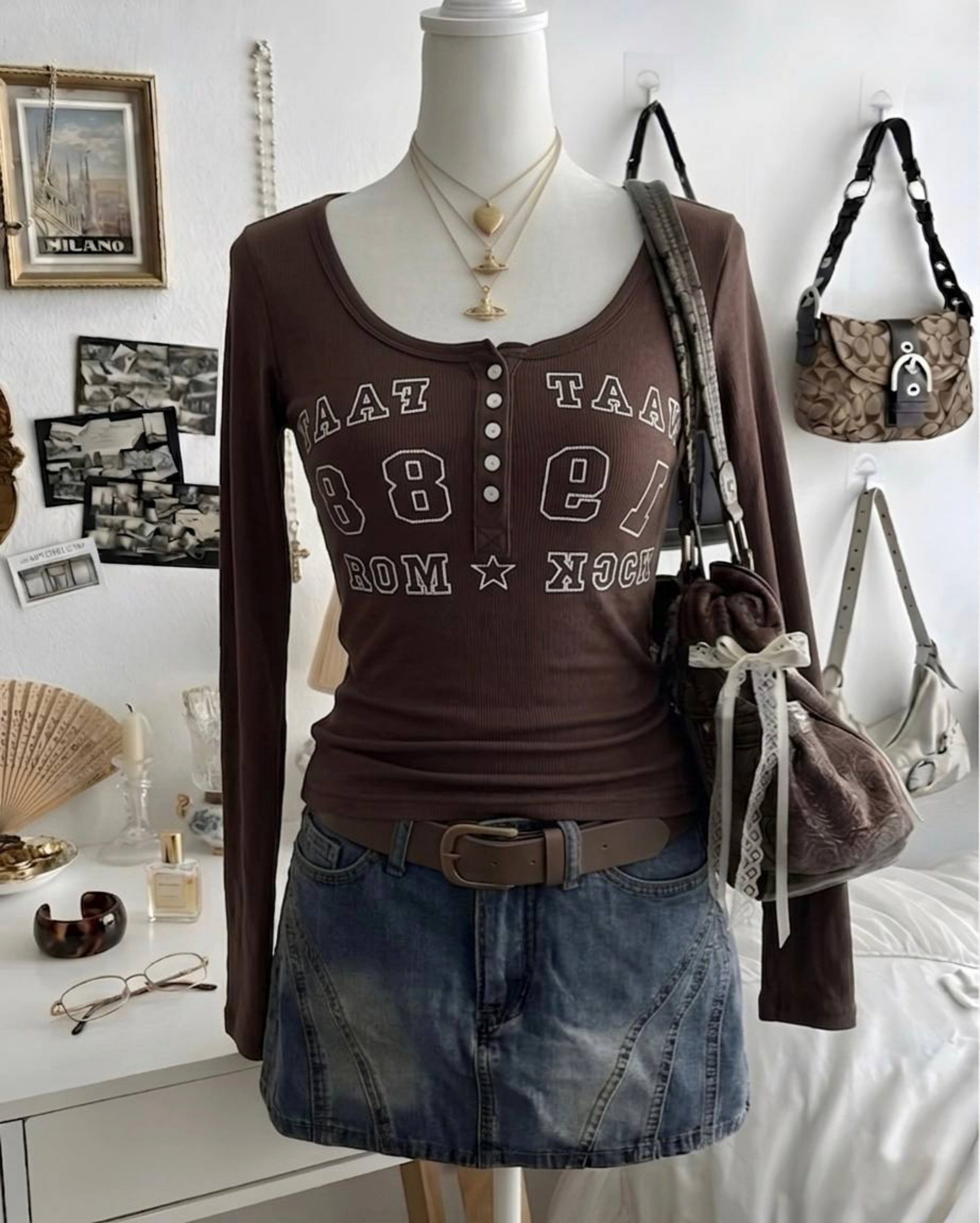 Mannequin wearing a brown long-sleeve shirt with text and a denim skirt, surrounded by bags and decor items.