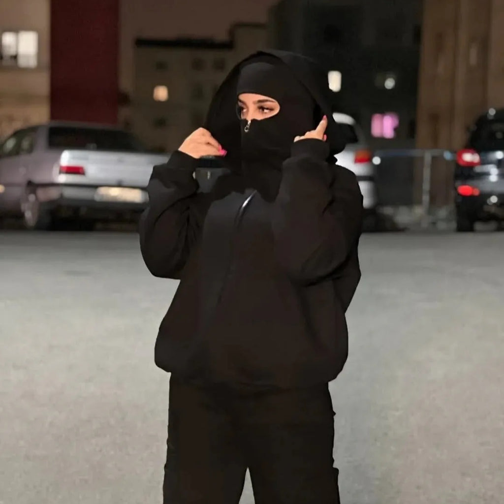 Person wearing a black hoodie and mask in an urban setting with cars and buildings in the background.