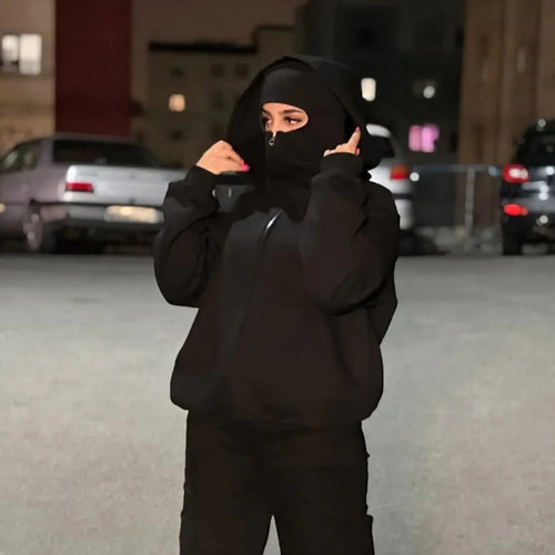 Person wearing a black hoodie and mask in an urban setting with cars and buildings in the background.