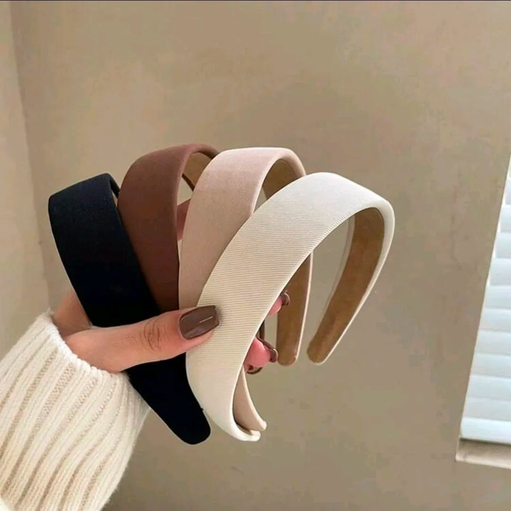 Four headbands in black, brown, beige, and white held by a hand against a neutral background.