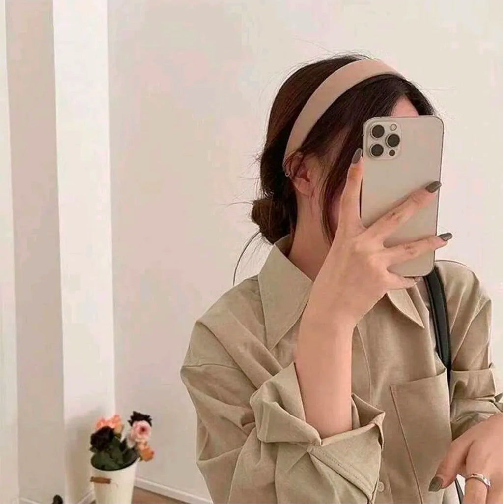 Person wearing a beige headband and holding a phone, standing against a plain wall.