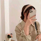 Person wearing a beige headband and holding a phone, standing against a plain wall.