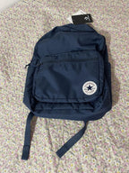 A navy blue Converse backpack with the classic circular white logo patch on the front pocket, resting on a pink and white floral-patterned fabric surface. A black hangtag is attached at the top.
