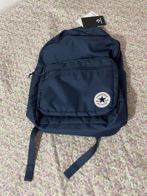 A navy blue Converse backpack with the classic circular white logo patch on the front pocket, resting on a pink and white floral-patterned fabric surface. A black hangtag is attached at the top.